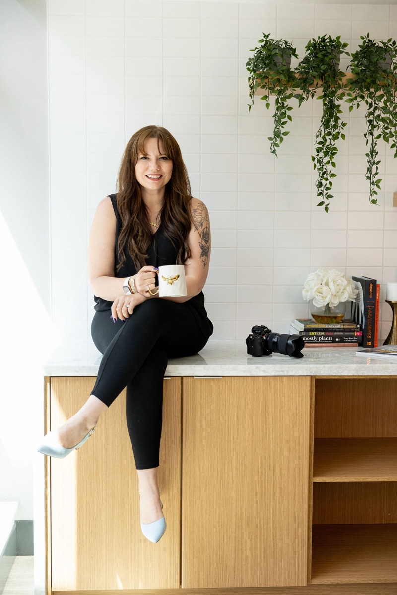 Brooke Redelle, host of HER., sitting on a counter with a coffee mug
